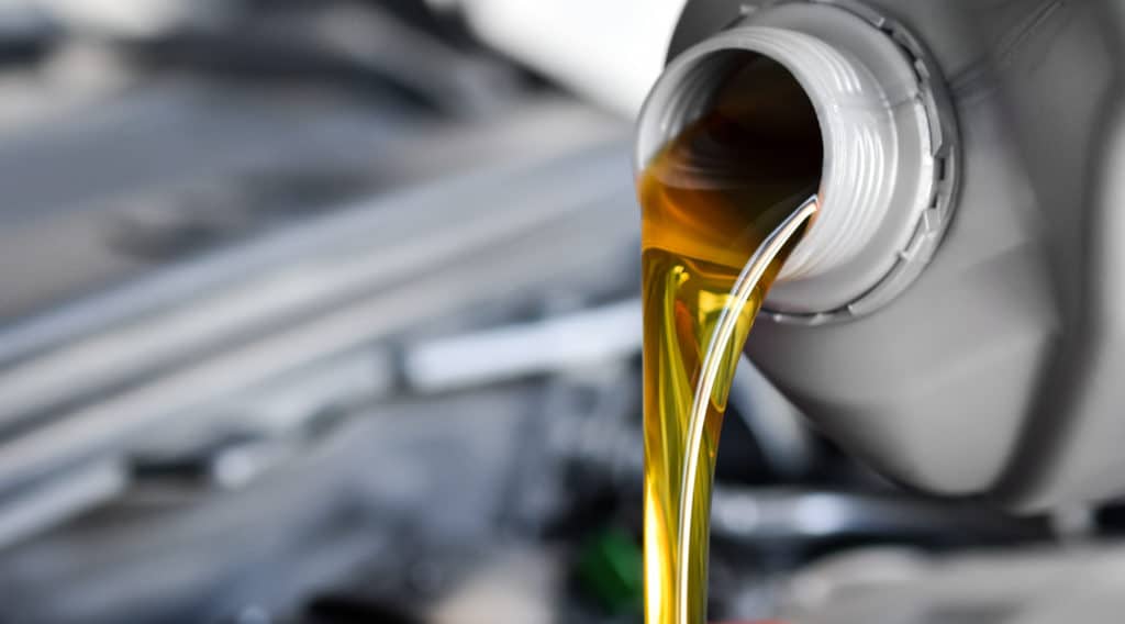 Das Bild zeigt, wie Motoröl aus einem grauen Kunststoffbehälter in einen Motor eingefüllt wird – eine typische Szene aus einer Autowerkstatt oder beim Ölwechsel. Das goldgelbe Öl ist klar und sauber, was auf frisches Öl hinweist.